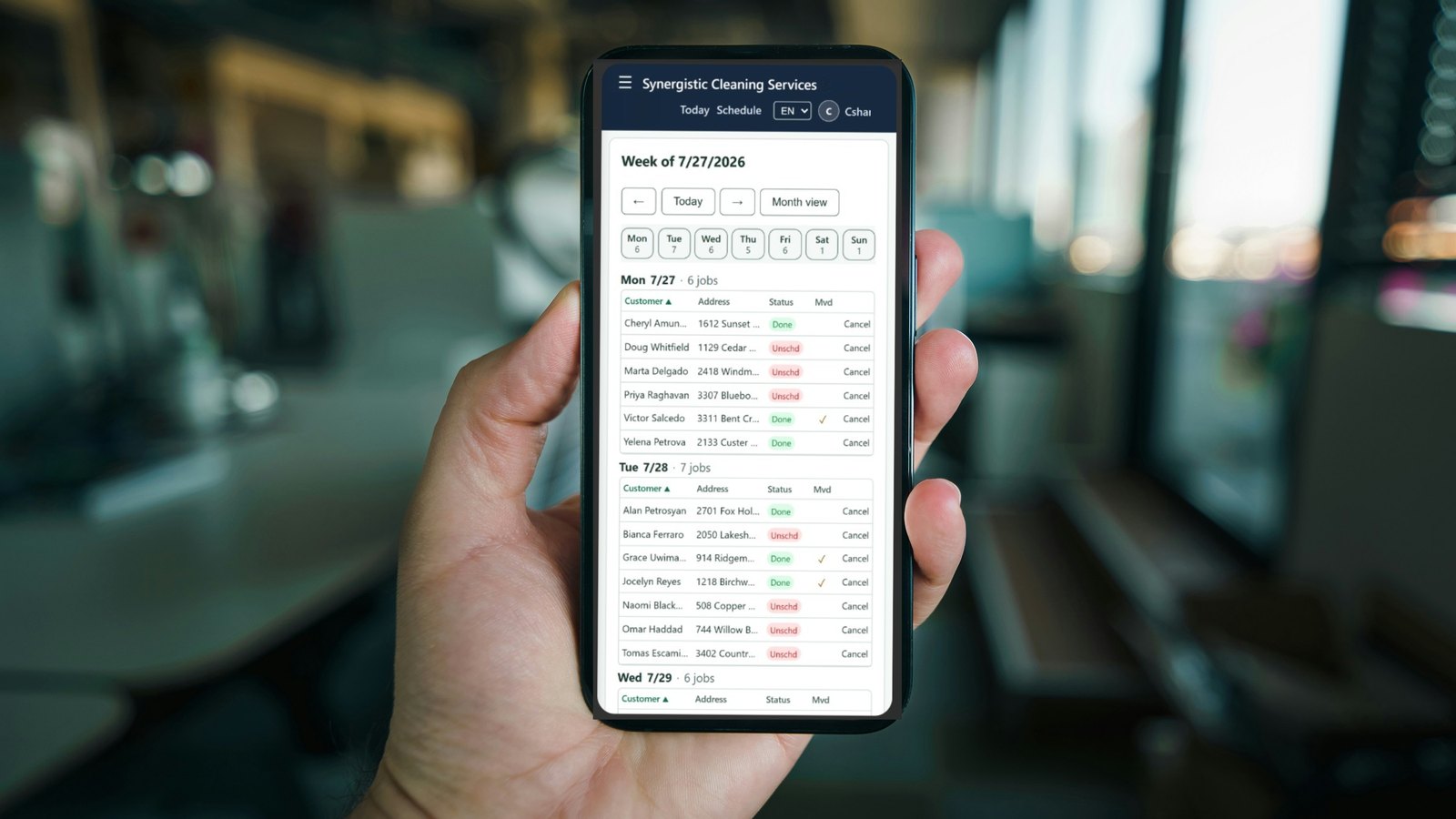 A hand holding a phone showing the week's schedule: each day a table of customers, addresses and job status